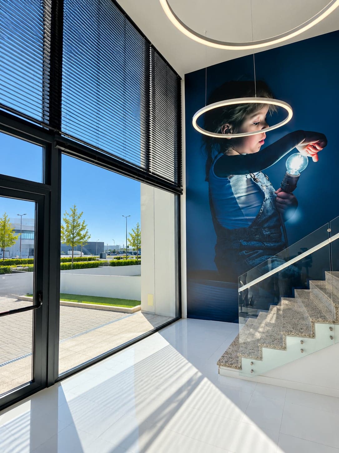 Hall com janela de grande altura e mural de uma criança com uma lâmpada, junto à escadaria